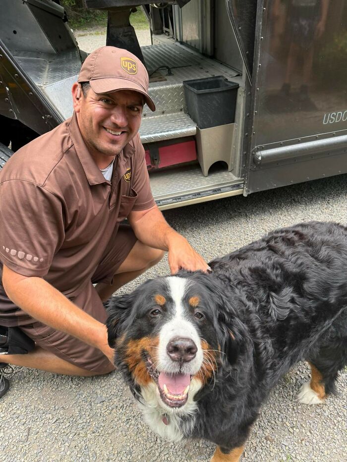 UPS driver smiling while petting a large friendly dog near a UPS delivery truck on a gravel driveway.