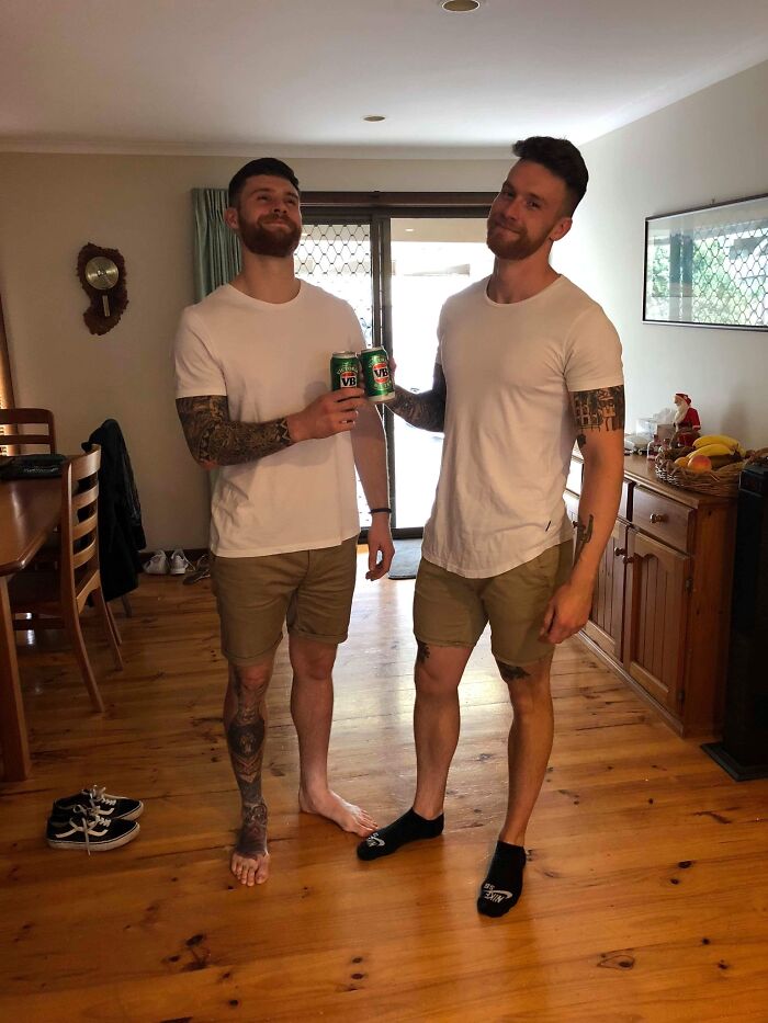 Two men with tattoos wearing white shirts and shorts toasting with cans, a wholesome moment indoors to distract from world horrors.