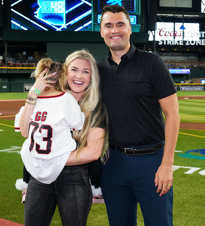 Charlie Kirk and his wife smiling on a baseball field with their child wearing a sports jersey during a family event.