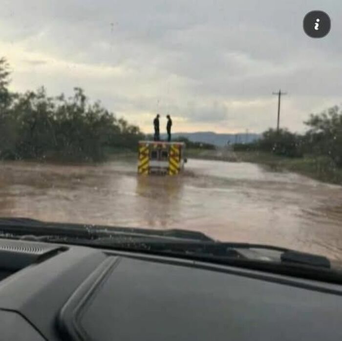EMT or paramedic standing on a vehicle surrounded by floodwaters on a rural road during overcast weather conditions.