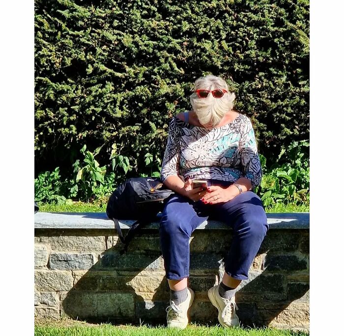 Woman sitting on stone bench, wearing sunglasses and colorful shirt, engrossed in phone in a funny street photo coincidence.