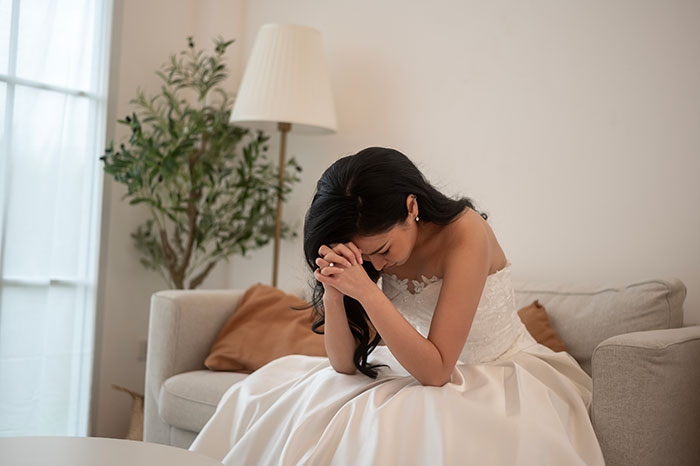 Bride in wedding dress sitting on couch with head bowed, showing distress over cheap wedding shower gifts controversy.