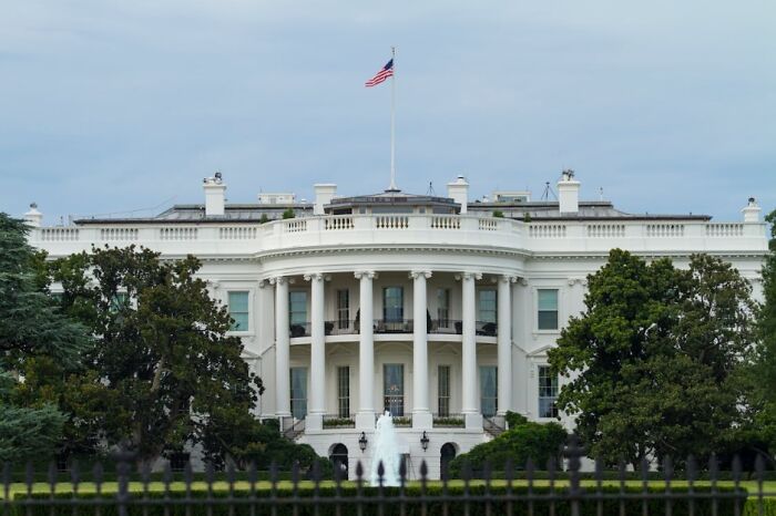 The White House exterior with American flag flying above, surrounded by trees and lawn under a cloudy sky.