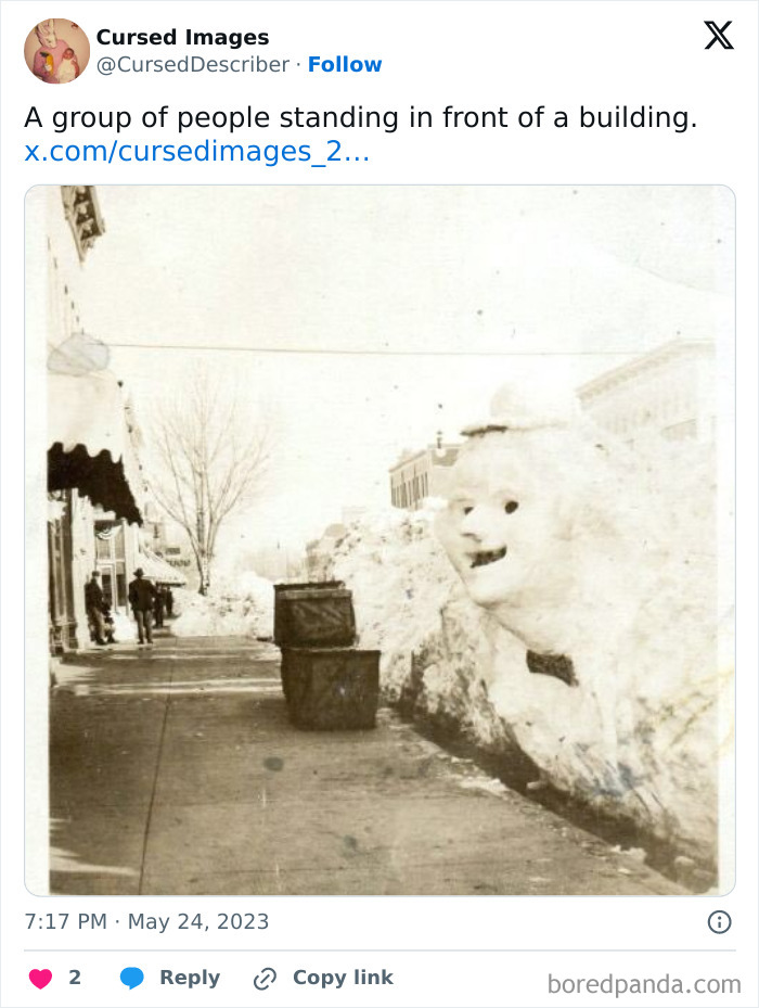 Creepy large snow sculpture with a distorted face on a snowy street next to trash bins in a vintage setting.