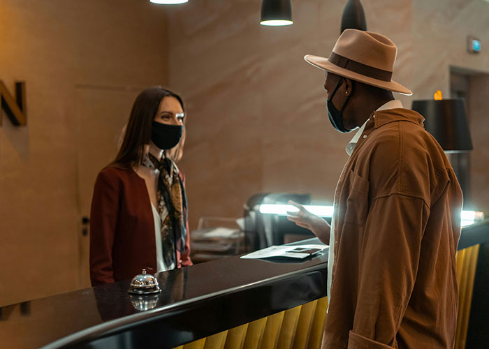 Man wearing a hat and mask talking to a masked receptionist at a hotel desk about incredible coincidences.