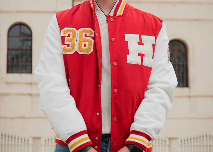 Person wearing red and white varsity jacket with 36 and H patches, standing outdoors near a building, scene of criminal fails.