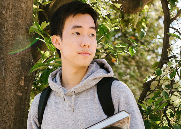 Young man in a gray hoodie standing by a tree outdoors, holding a book, illustrating incredible coincidences that sound fake.