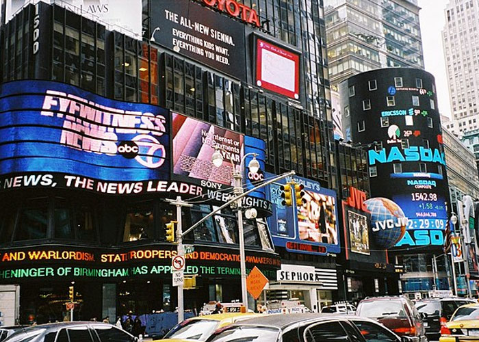 Busy urban street with electronic billboards and news displays highlighting societal impact of historical events.
