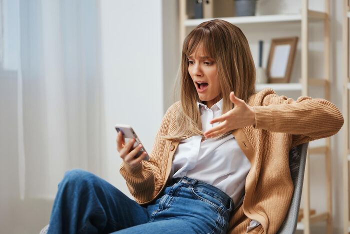 Woman sitting in a chair looking shocked and upset at her phone, highlighting absurd dealbreakers in relationships.