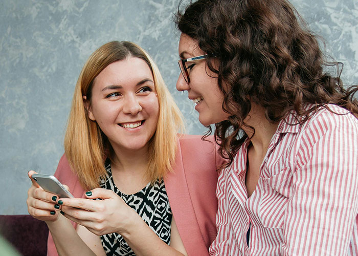 Two women smiling and chatting closely while one holds a smartphone, highlighting creepy behaviors that cross the line.