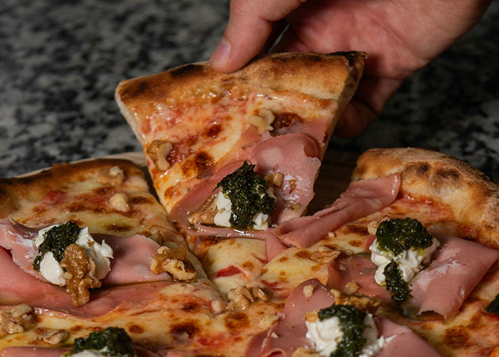 Hand picking a slice of gourmet pizza topped with ham, walnuts, cheese, and pesto, highlighting common purchases.