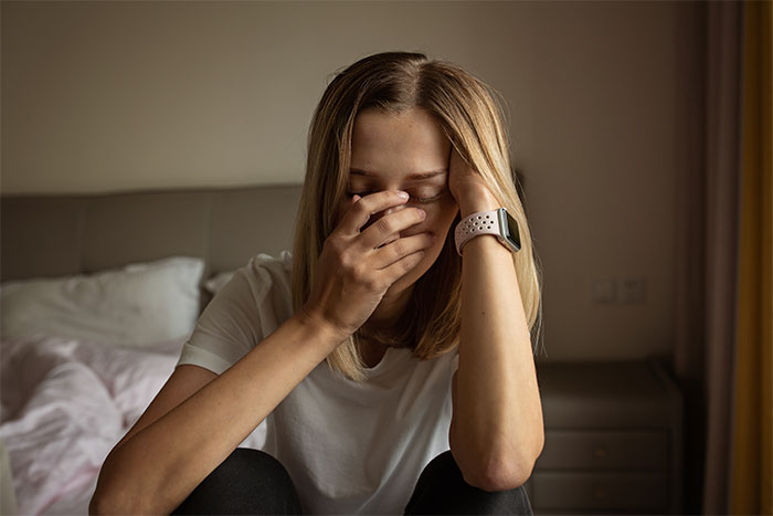 Teen girl looking disappointed and deeply hurt sitting on bed wearing a white shirt and smartwatch at home.