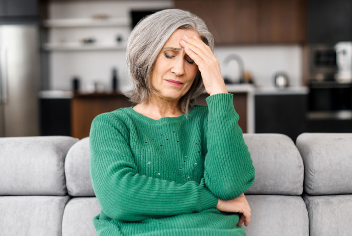 Mature woman in green sweater looking stressed and upset while sitting on a couch, struggling with sibling&rsquo;s engagement news.