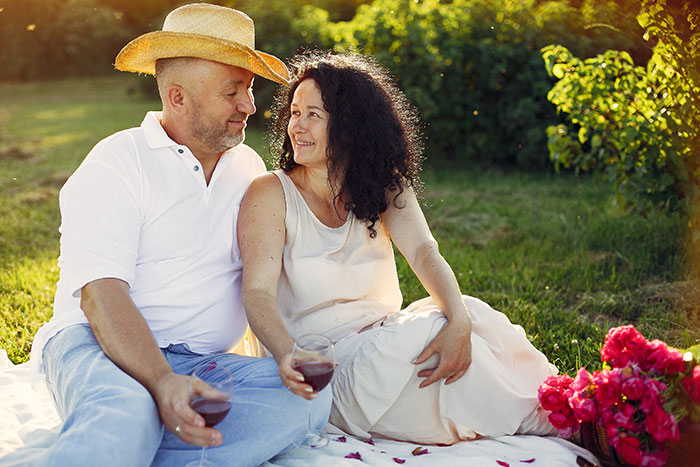 Couple sitting on a picnic blanket outdoors enjoying wine, with sunlight and greenery in the background on a calm day.