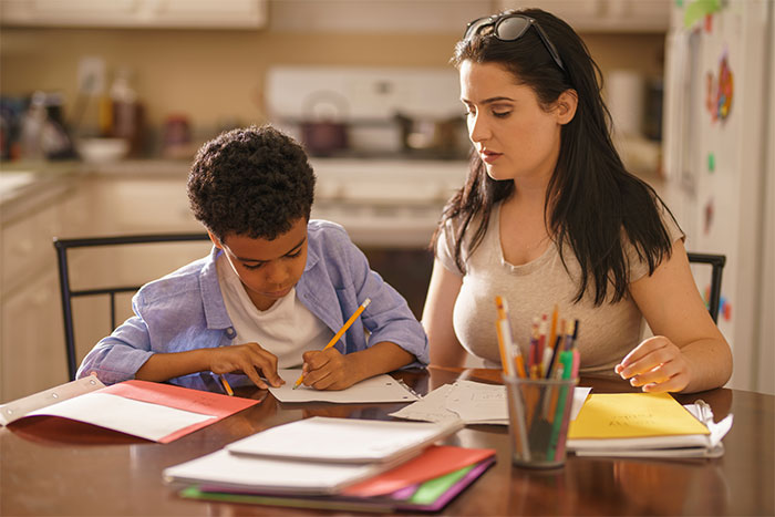 Boy working on homework at kitchen table while babysitter supervises in a home setting with school supplies nearby