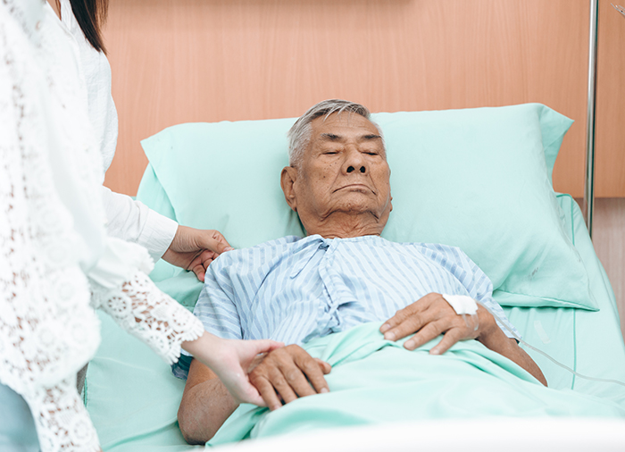 Elderly man resting in hospital bed while woman holds his hand, reflecting emotional wedding gift and family conflict topic.