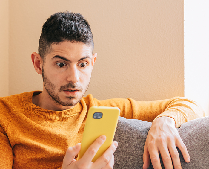 Man wearing orange sweater looking shocked at phone while sitting on a couch, depicting relationship trust issues.