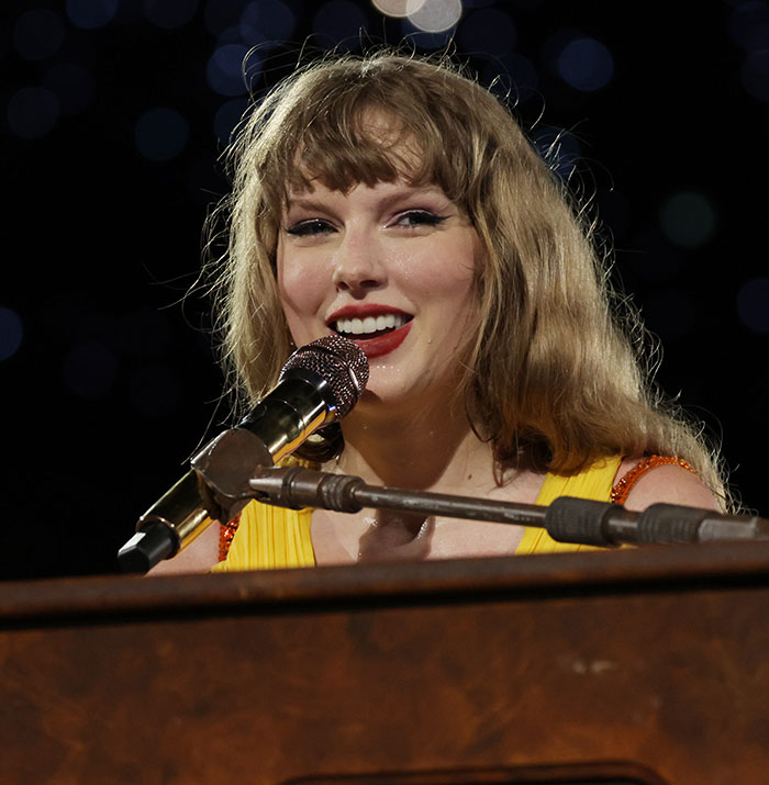 Taylor Swift singing at a piano during a live performance with a focused audience in the background.