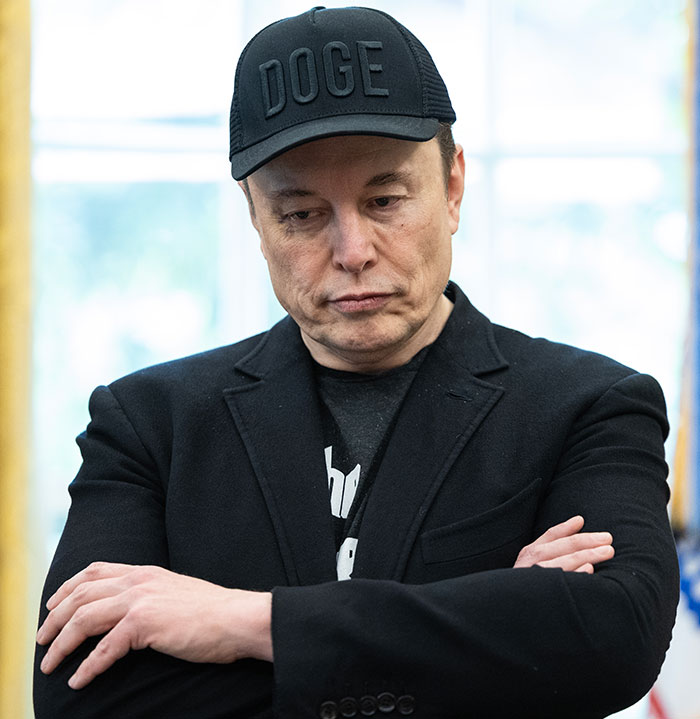 Elon Musk wearing a black DOGE cap and blazer, looking thoughtful with arms crossed in an indoor setting. Elon Musk wearing a black DOGE cap and blazer, looking thoughtful with arms crossed in an indoor setting.