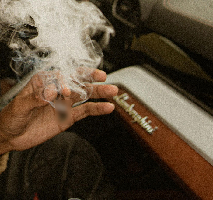 Hand holding a smoldering cigarette inside a car with smoke rising, related to hotboxing and child endangerment.