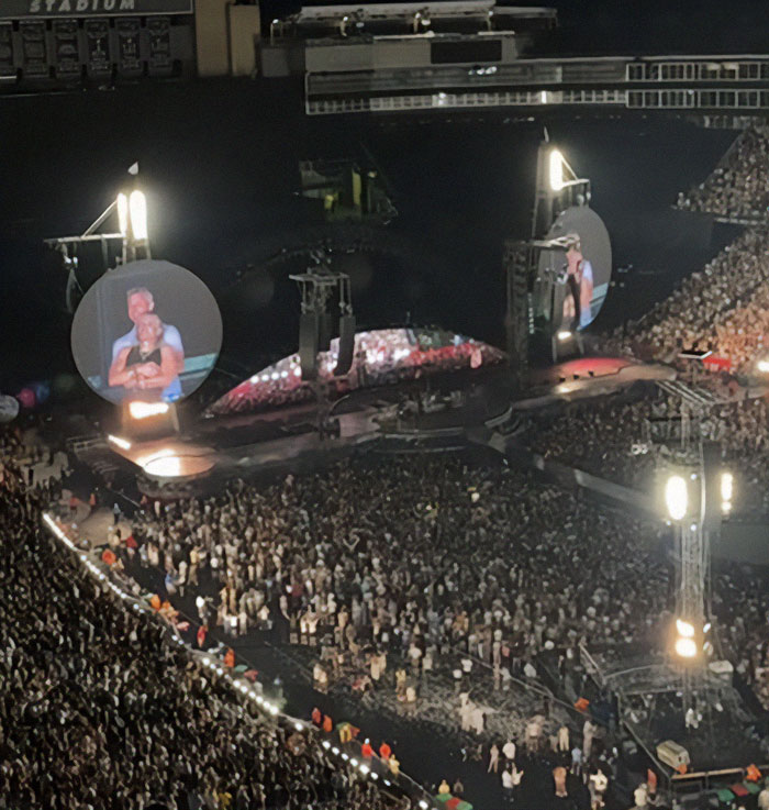 Large concert crowd at Coldplay stadium show featuring a kiss cam moment on giant circular screens.