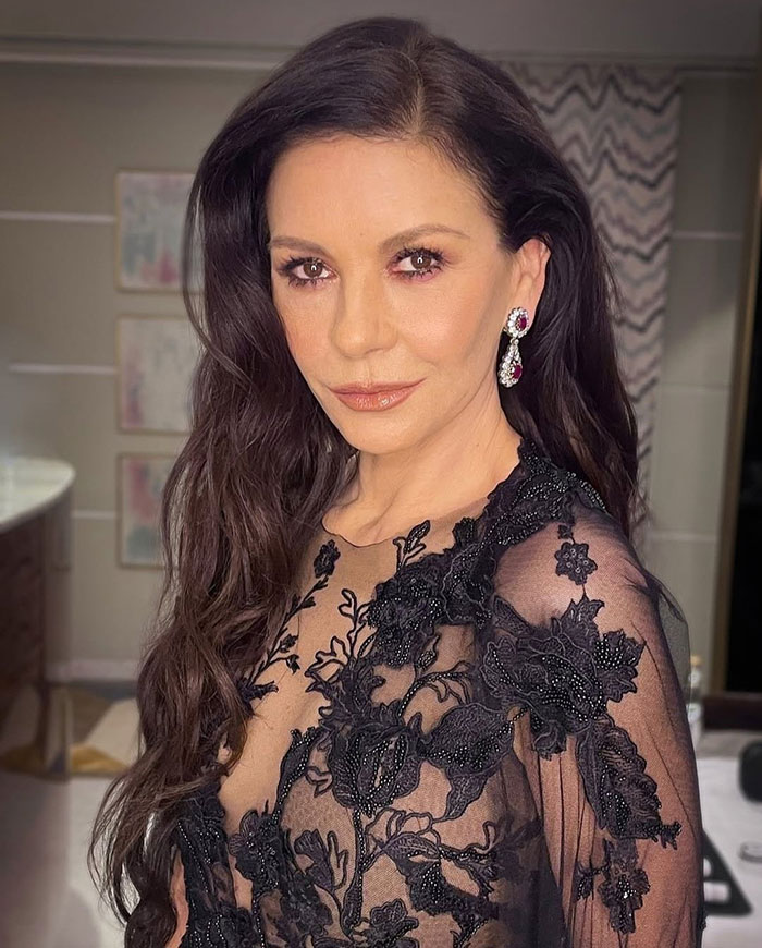 Catherine Zeta-Jones posing in a black floral lace dress and statement earrings with long wavy hair indoors.