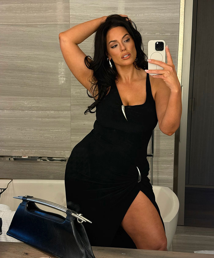 Ashley Graham wearing a body-hugging black dress revealing her figure, taking a mirror selfie in a modern bathroom.
