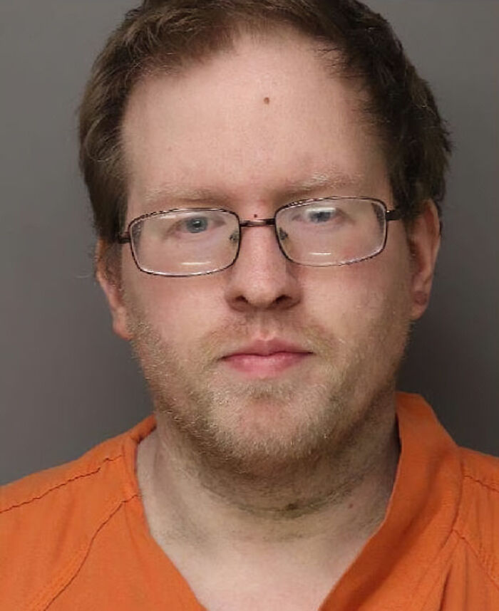 Mugshot of pastor's son wearing glasses and orange prison uniform, accused of disturbing acts against four captive adults.