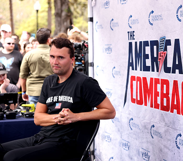 A man sitting near a banner with The American Comeback logo, with people and cameras in the background at an event. A man sitting near a banner with The American Comeback logo, with people and cameras in the background at an event.