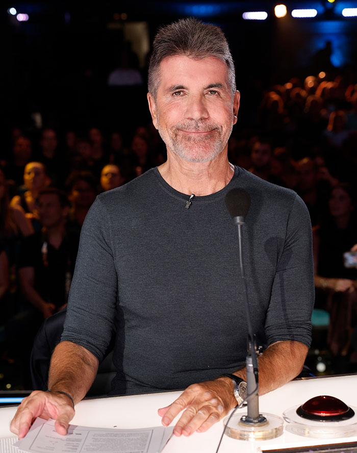 Simon Cowell sitting at a judge&rsquo;s panel, smiling, during a televised talent show with audience in the background.