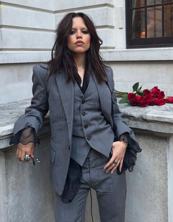 Jenna Ortega in a gray pinstripe suit, posing outdoors with a bouquet of red roses, styled by her Emmy outfit stylist.