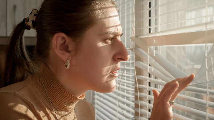 Woman peering through window blinds with concern, anxious about a stalker customer who rang the doorbell unexpectedly.