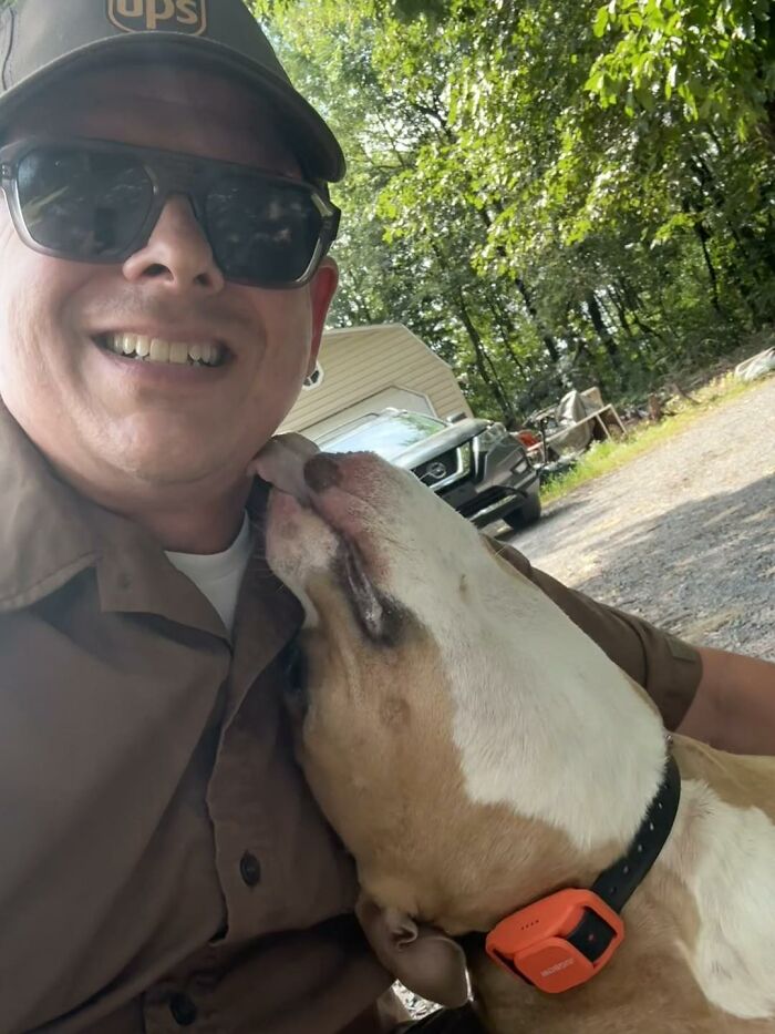 UPS driver smiling with sunglasses while an adorable dog wearing a collar affectionately licks his face outdoors.