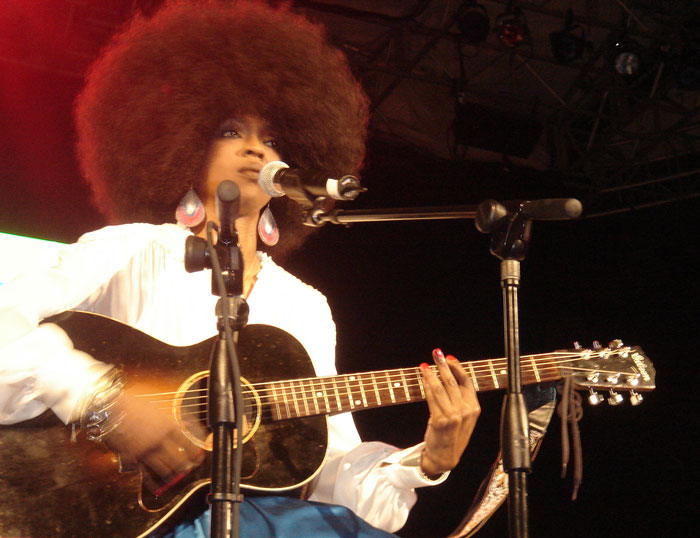 Female musician with an afro playing an acoustic guitar and singing on stage during a live concert performance.