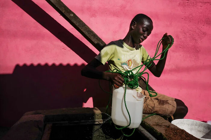 Child of the world sitting by a pink wall, holding a rope and water container in powerful photograph by Andrea Torrei.