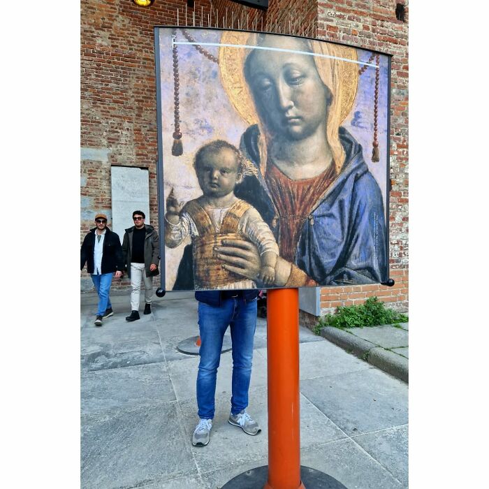 Person standing behind street photo display creates a funny coincidence with religious painting, blending art and reality.