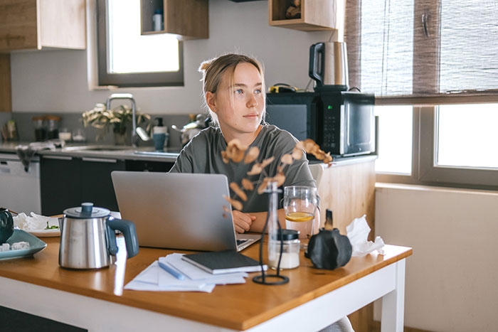 Young woman working on laptop at home, reflecting on inclusion of AI into everything in a modern kitchen setting.
