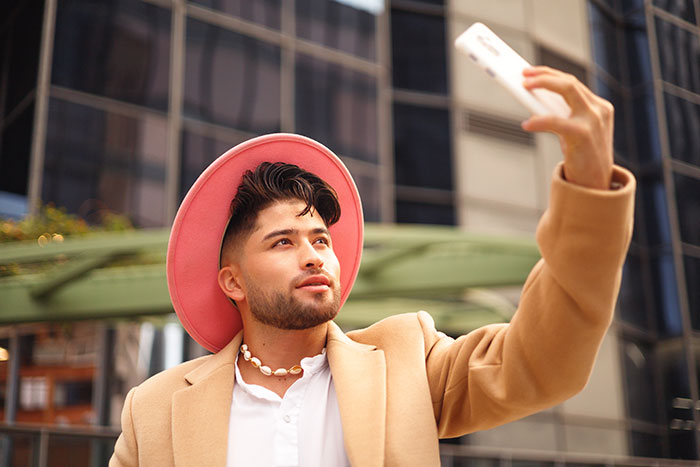 Young man wearing a pink hat and beige coat taking a selfie outdoors, highlighting inclusion of AI into modern trends.