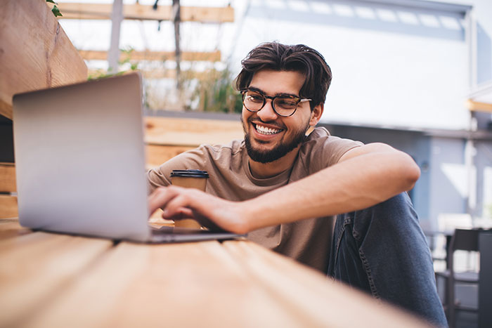 Young man smiling while using laptop outdoors, illustrating the inclusion of AI into everything in modern trends.