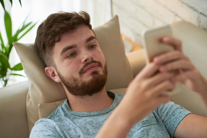 Young man surfing online on his smartphone discovering his partner's secret stories while relaxing on a couch at home.