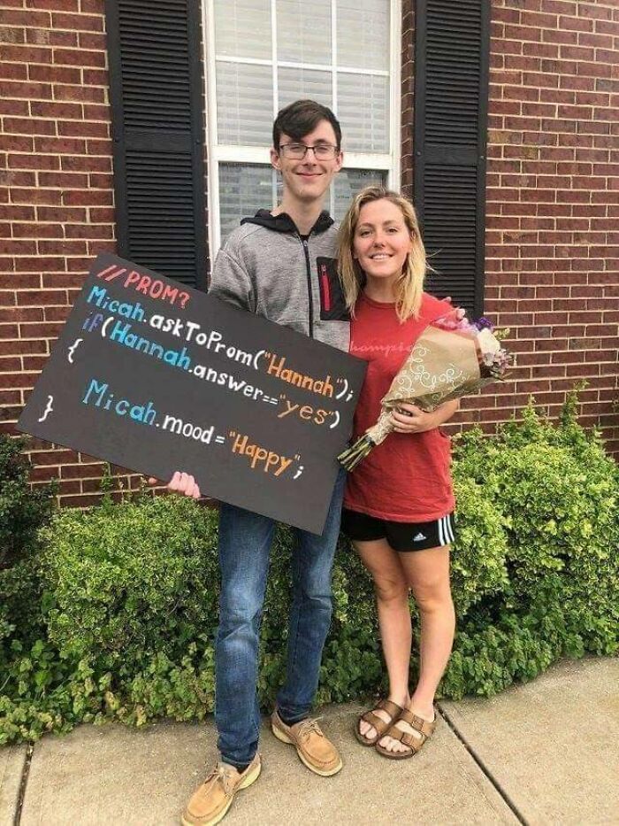 Young man holding a funny programming meme promposal sign standing next to a woman with flowers outside a brick house.
