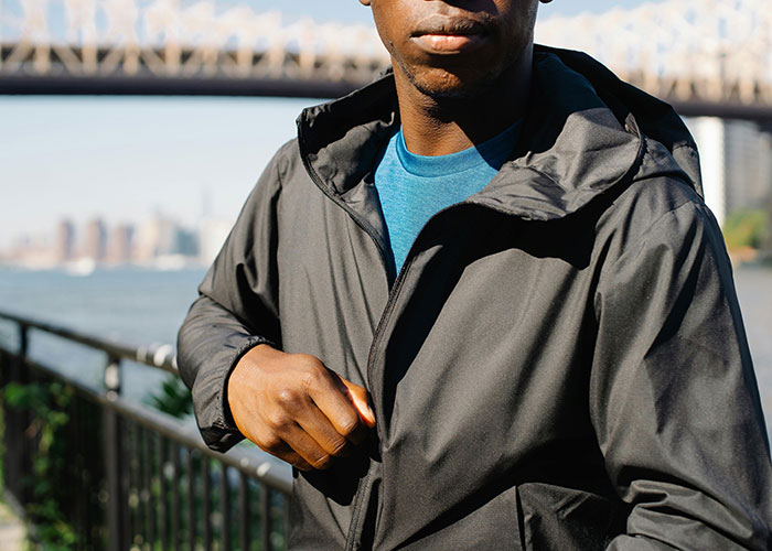 Man in a black jacket attempting to hide while standing near a waterfront railing in a cityscape background, criminal fails theme