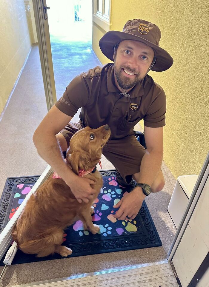 UPS driver in uniform kneeling and smiling while petting an adorable dog on a colorful paw print mat at a doorstep