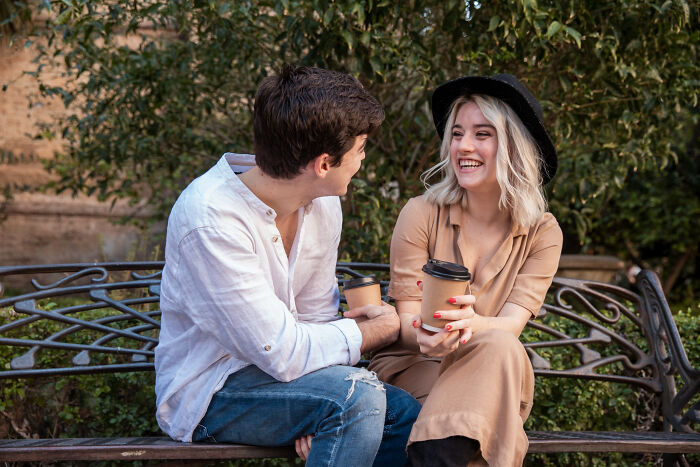 Young couple sitting on a park bench, smiling and chatting while holding coffee cups, illustrating partners' secret stories.