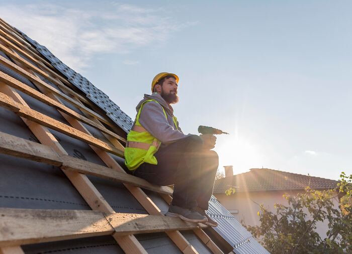 Man in safety vest and helmet sitting on roof holding drill, reflecting on moments of falling out of love, with sunset backdrop.