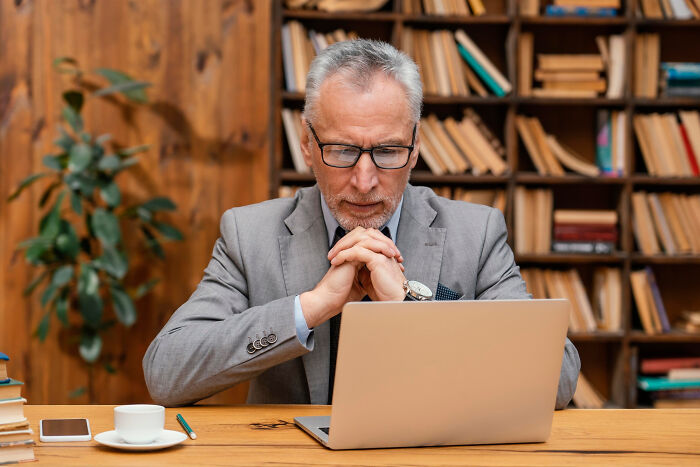 Older man in glasses and gray suit having a mind-boggling conversation on laptop in a cozy library setting.