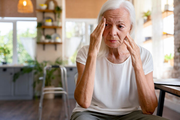 Elderly woman sitting indoors with a pained expression, touching her temples, showing signs of distress or worry.