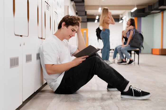 Young man sitting by lockers, reading a book and looking thoughtful, illustrating discovery of secret stories online.