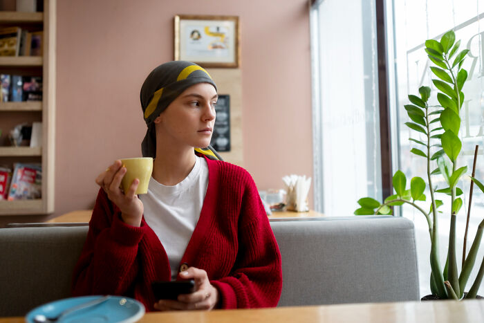 Person in a red sweater holding a cup and phone, looking out the window while surfing online at a cafe.
