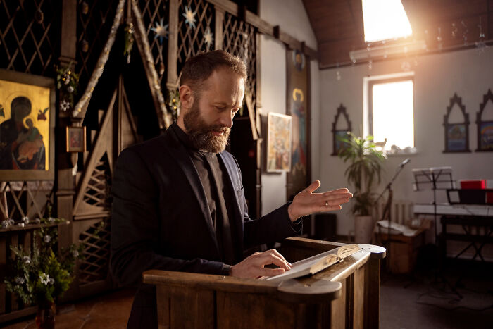 Man in a black suit reading and revealing shocking family secrets inside a warmly lit, decorated room with religious icons.